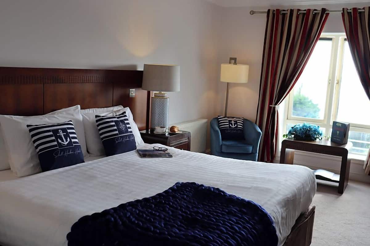 Habitación de hotel con una cama grande, una mesita de noche con lámpara, una silla y vistas al exterior a través de una ventana con cortinas.