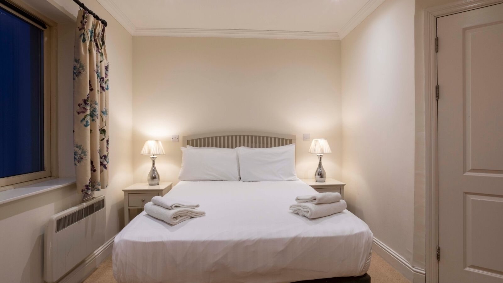 Un lit bien fait, avec du linge de lit blanc, deux lampes de chevet et une fenêtre ornée de rideaux à motifs floraux.