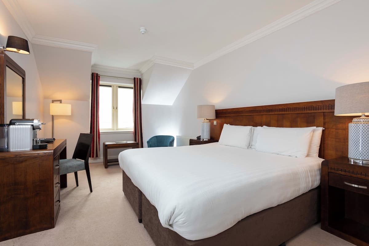 Habitación de hotel con una cama grande, un escritorio con silla, una lámpara y un ventanal con cortinas.