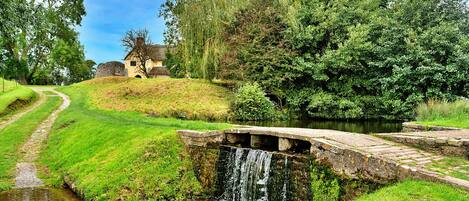 A stone bridge over a small stream with a house in the background.