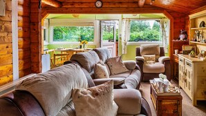 A cozy log cabin interior with a wooden ceiling, a sofa with cushions, a dining table with chairs, and a view of greenery outside. - Mountain View (Trawsfynydd)