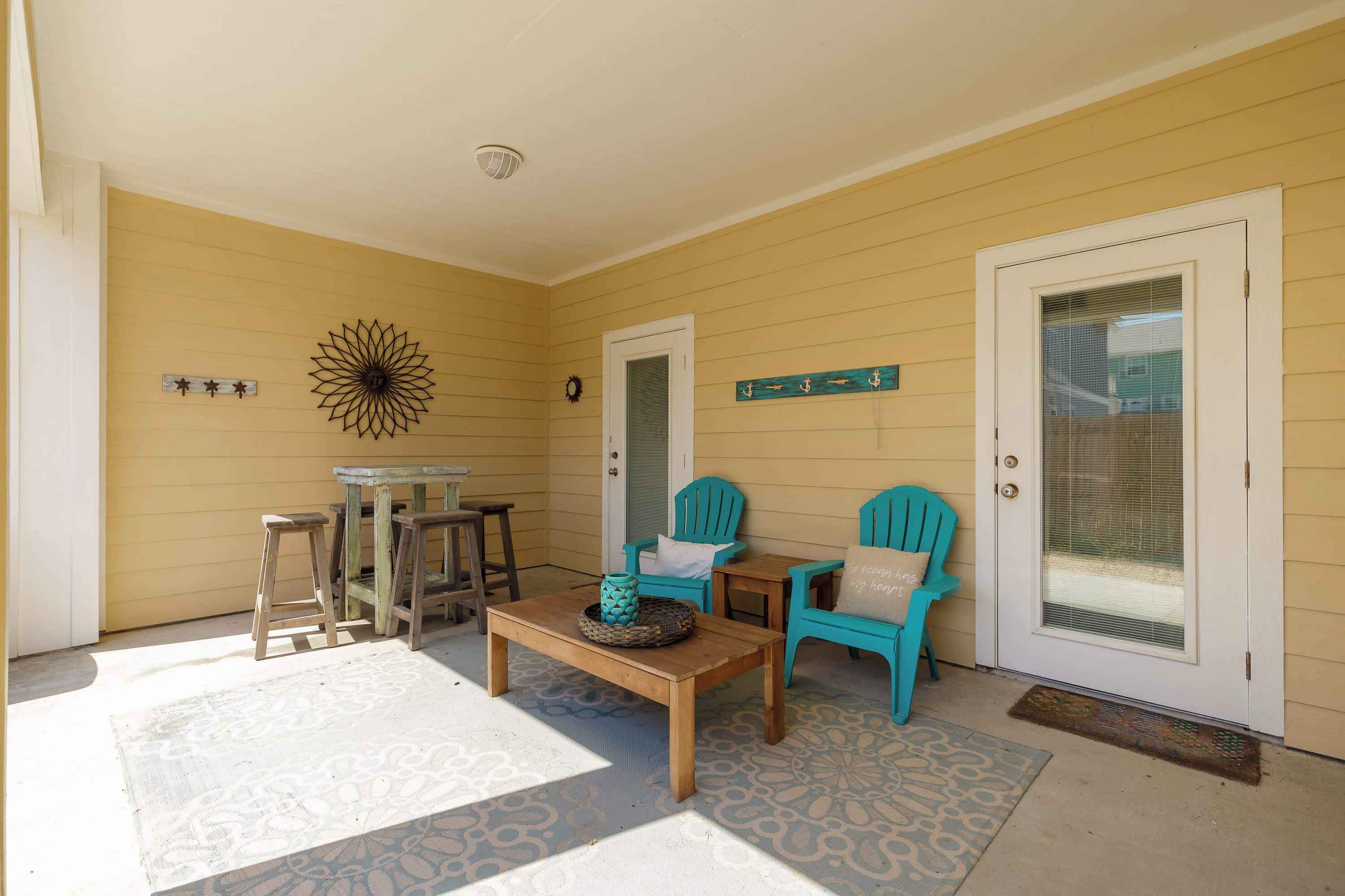 A covered porch with a wooden table, two turquoise chairs, and a decorative wall piece.