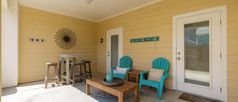 A covered porch with a wooden table, two turquoise chairs, and a decorative wall piece.