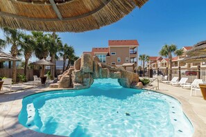 A swimming pool with a rock arch, surrounded by lounge chairs and palm trees, with buildings in the background. - Fun 4 Bedroom Resort Pool Sleeps 12 (Port Aransas)