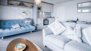 Una sala de estar moderna con un sofá y una mesa de centro, una cocina con armarios blancos y un horno empotrado, y un candelabro.