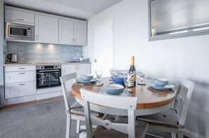 Uma cozinha moderna com armários brancos, micro-ondas e forno. Há uma mesa de jantar com tigelas azuis e copos de vinho.