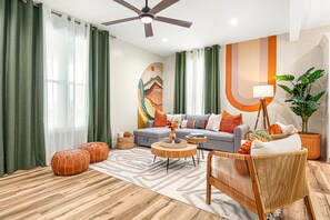 Uma sala de estar moderna com sofá, pufes, mesa de centro, poltrona de vime e uma grande pintura na parede.