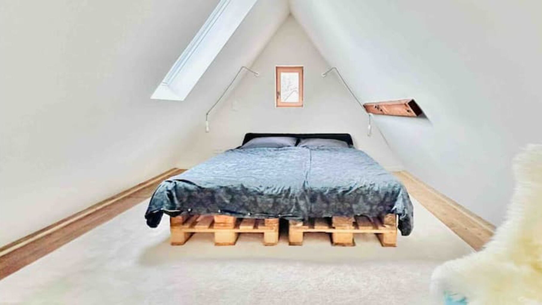Un pequeño dormitorio en ático, con forma de triángulo, que tiene una cama sobre una base de madera, un tragaluz y un cuadro en la pared.