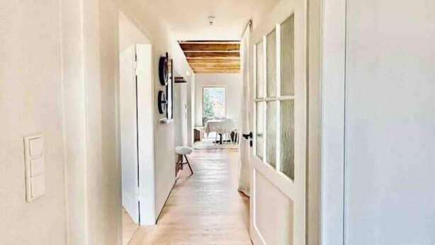A hallway with wooden flooring and white walls, leading to a room with a large window and a ceiling with exposed beams. - Galeriewohnung mit Ambiente (Wertheim)