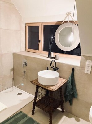 Une salle de bain avec un miroir rond, un lavabo blanc posé sur un tabouret en bois, une serviette accrochée au mur et un espace douche.