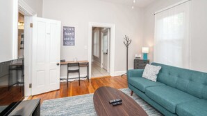 Un salon avec un canapé de couleur turquoise, une table basse, un téléviseur, un bureau avec une chaise et un panneau sur lequel on peut lire : « Travaillez dur et soyez aimable ».