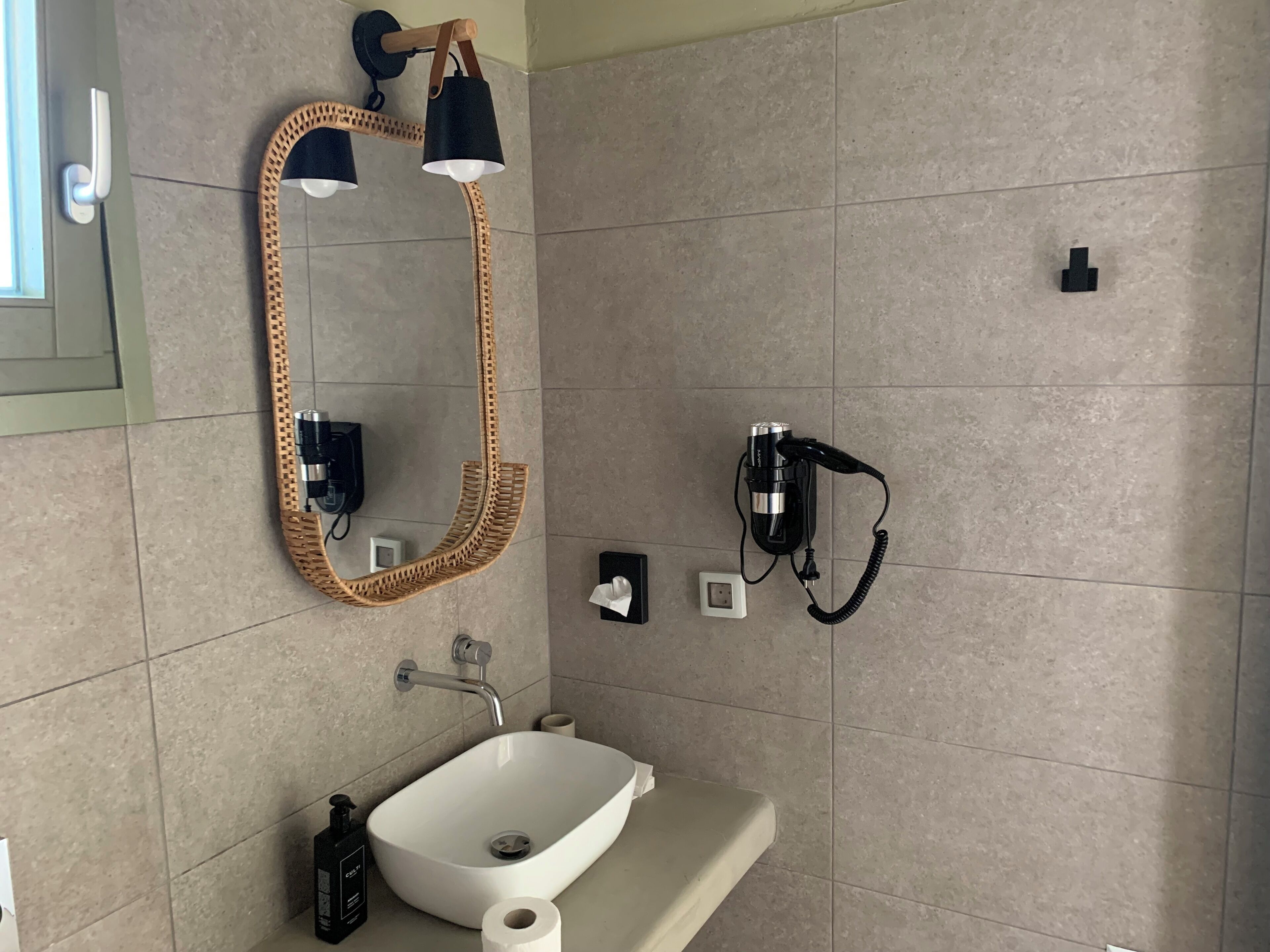 A bathroom with a white sink, a mirror with a woven frame, a black telephone, and a towel dispenser.