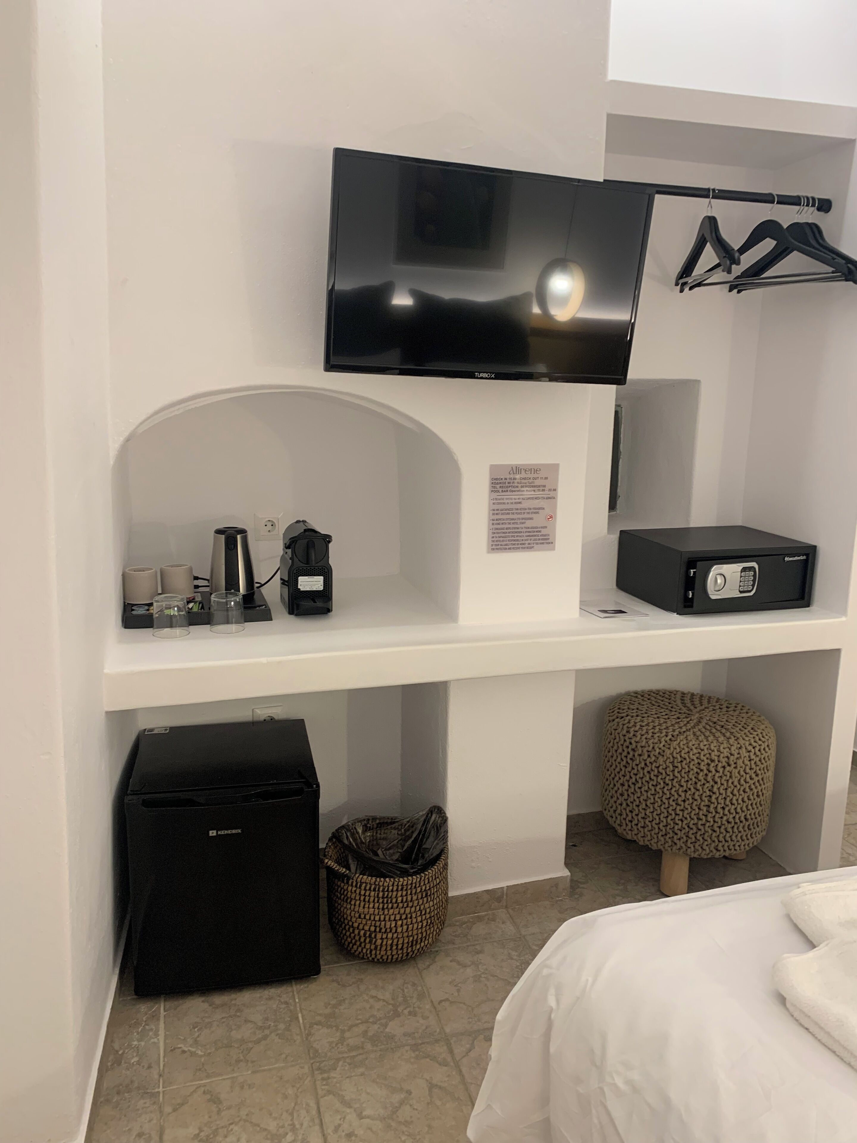 Hotelkamer met een flatscreen televisie bevestigd boven een witte console, een minibar, een zwarte koffer, een geweven pouf en een bed met wit beddengoed.