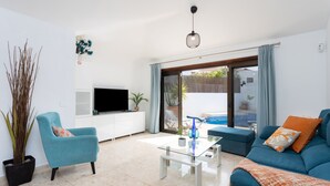 Una sala moderna con un sofá, un sillón y una mesa de centro. Se observa un televisor de pantalla plana sobre un mueble blanco, y una planta en maceta.
