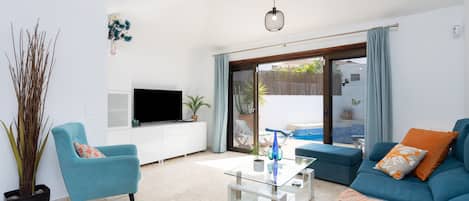 Una sala moderna con un sofá, un sillón y una mesa de centro. Se observa un televisor de pantalla plana sobre un mueble blanco, y una planta en maceta.