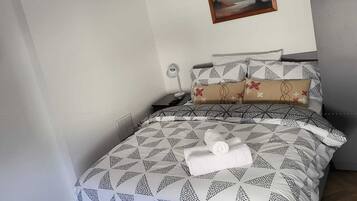 A neatly made bed with a geometric patterned bedspread, a painting of trees on the wall, and a wooden floor.