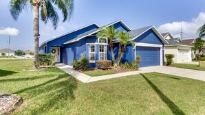 A single-story house with a blue exterior, a garage, and a front yard with a palm tree and shrubs. - Davenport Home w/ Game Room, Home Theater, Pool (Davenport)