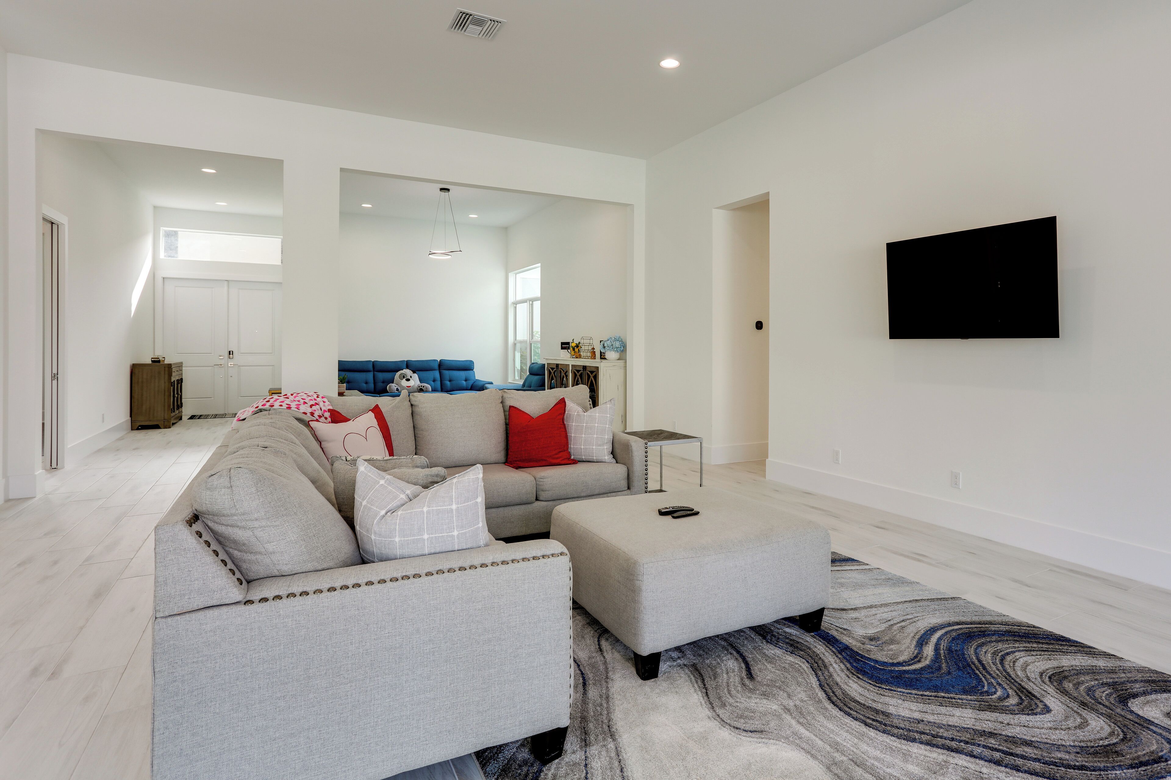 A modern living room with a grey sectional sofa, a matching ottoman, and a large flat-screen TV mounted on the wall.
