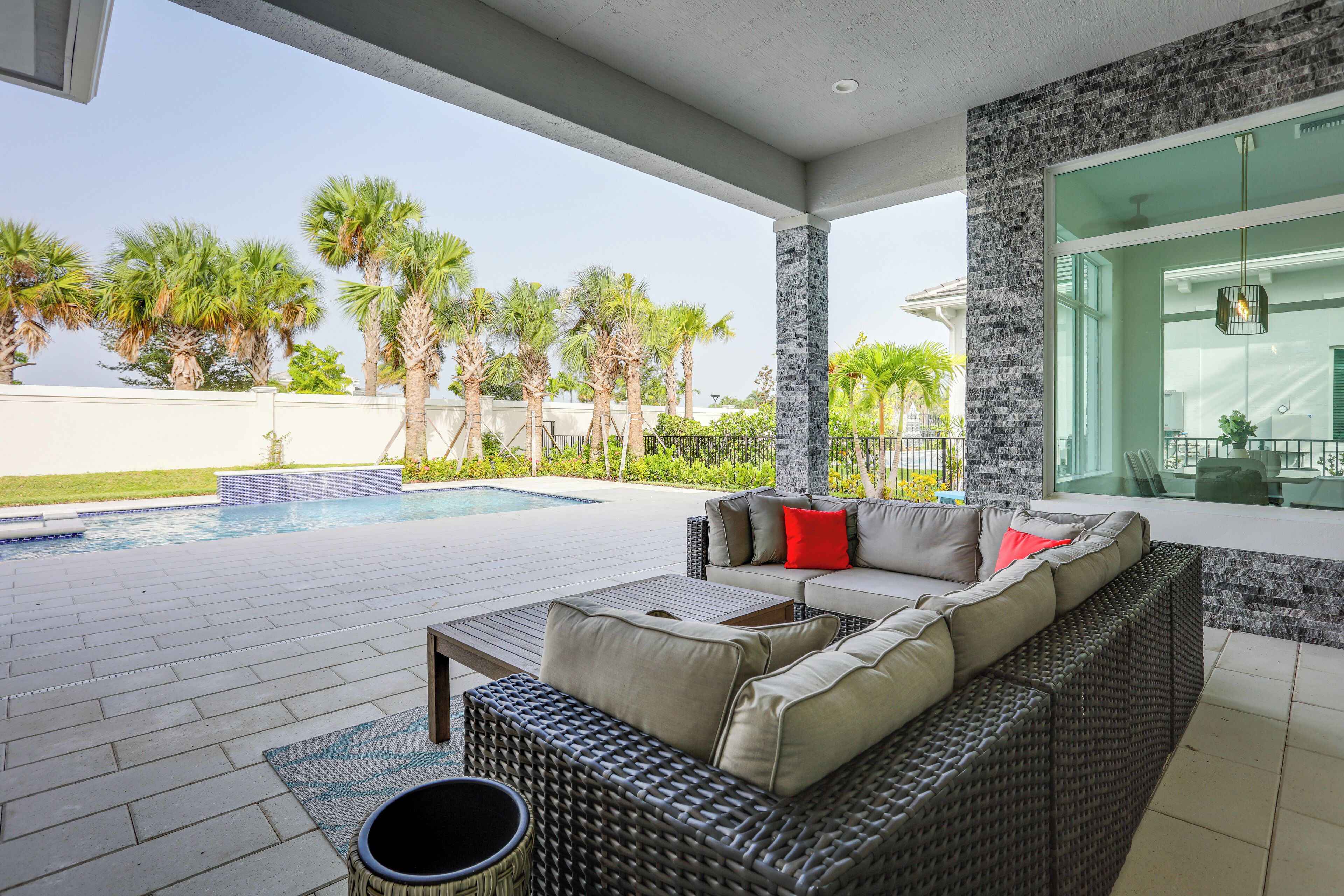 A covered outdoor area with a wicker sofa, a pool, and palm trees.
