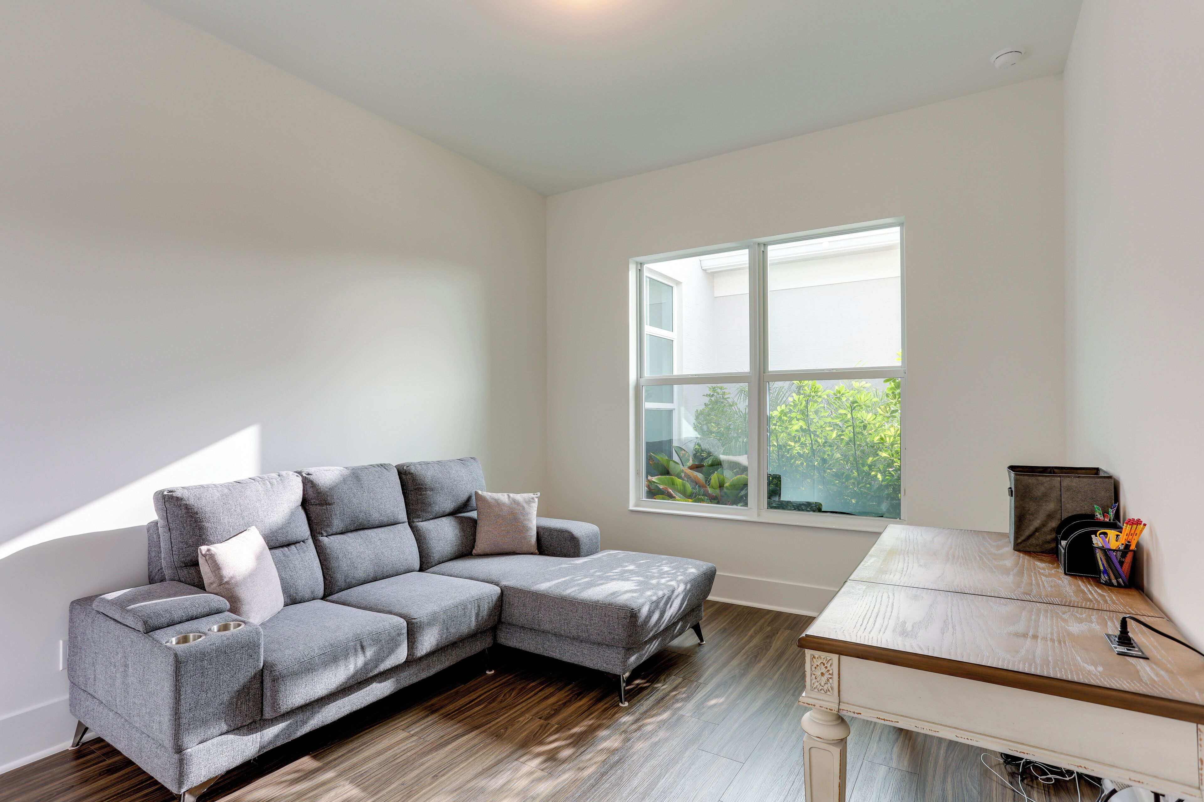 A modern living room with a grey sectional sofa, a wooden coffee table, and a large window providing a view of greenery outside.