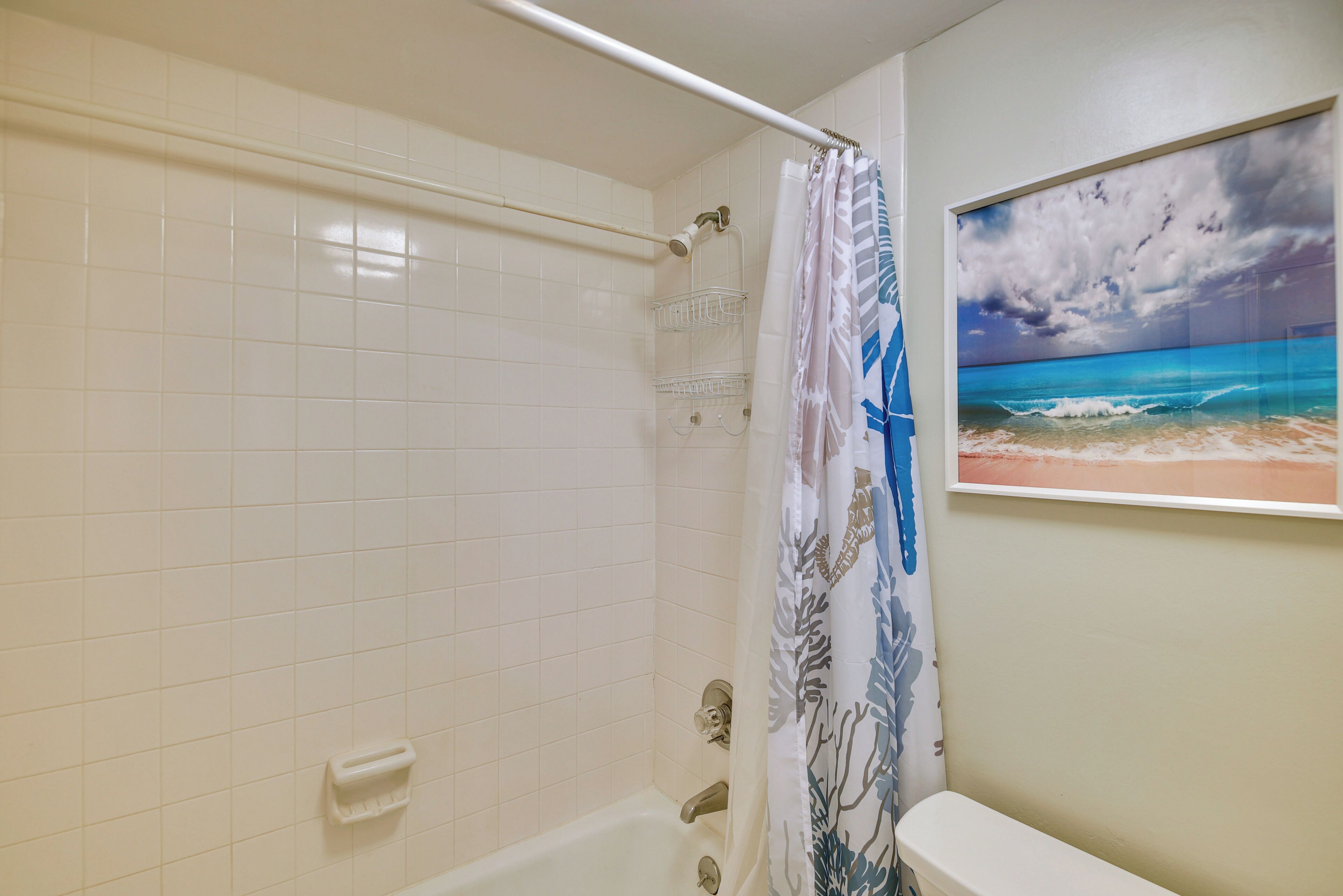 Un baño con paredes de azulejos blancos, una bañera, una cortina de ducha con diseño azul y blanco, y un cuadro en la pared que muestra una playa.