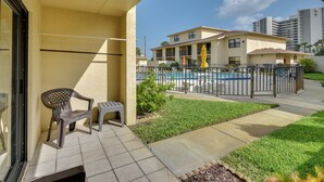 A balcony with a chair and a small table, a pool area with a fence, and a building in the background. - Daytona Beach Shores Condo Rental: Walk to Beach! (Daytona Beach)