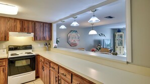 A kitchen with wooden cabinets, a white countertop, and a stove. There is a window with a view of a dining area and a cabinet with decorative items. - Daytona Beach Shores Condo Rental: Walk to Beach! (Daytona Beach)