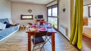 A cozy room with a wooden dining table set for four, a sofa, a TV, and a window with a view of the outdoors. - Cosy apartment for 6 people with WIFI, pool and TV (Le Corbier)