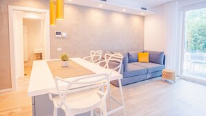 A modern living room with a dining table, white chairs, a grey sofa with a yellow pillow, and a window with a view of greenery. - Garda Luxury Suite - One Bedroom Apartment, Sleeps 2 (Padenghe sul Garda)
