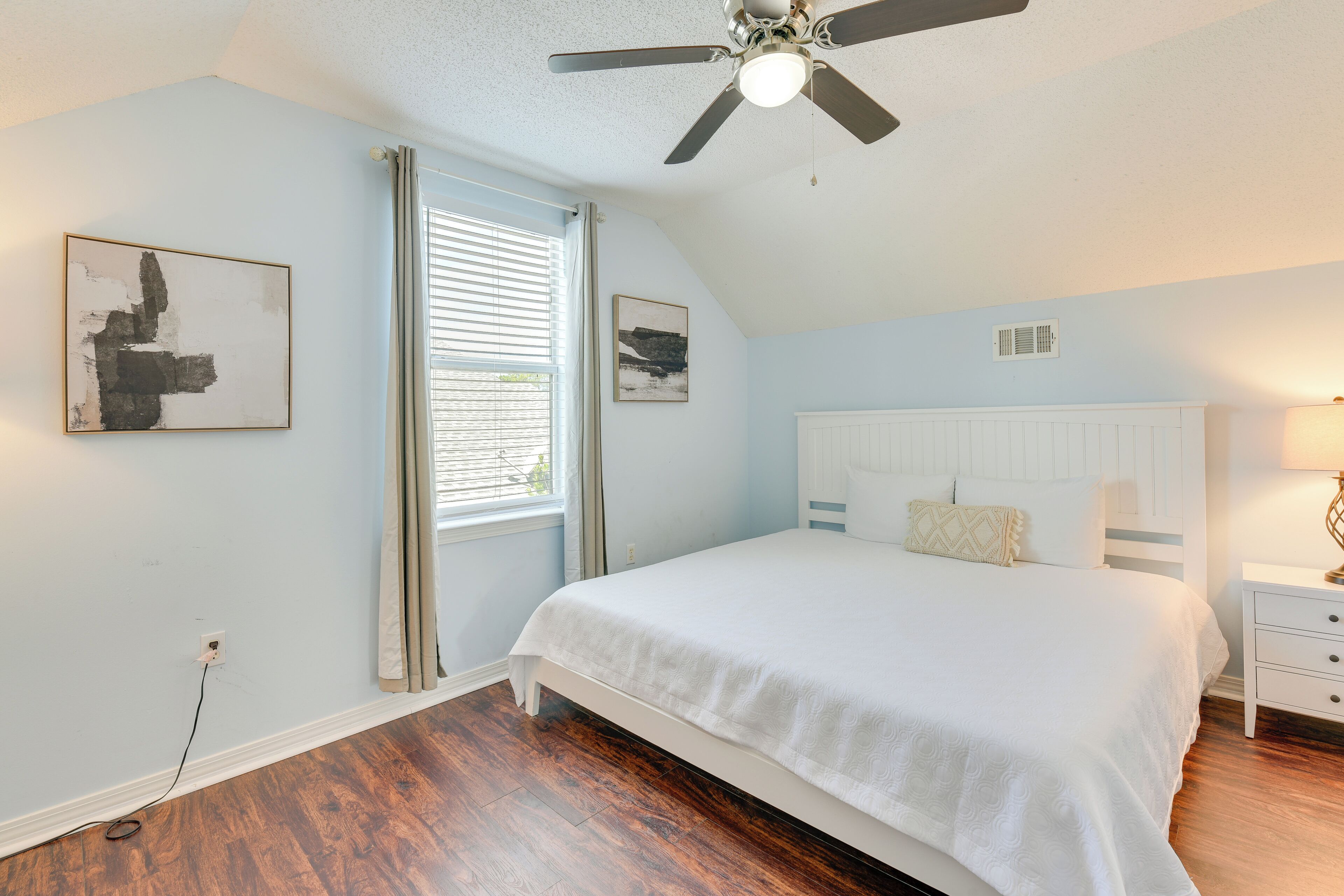 A bedroom with a white bed, a ceiling fan, a window with blinds, and a wall-mounted picture.