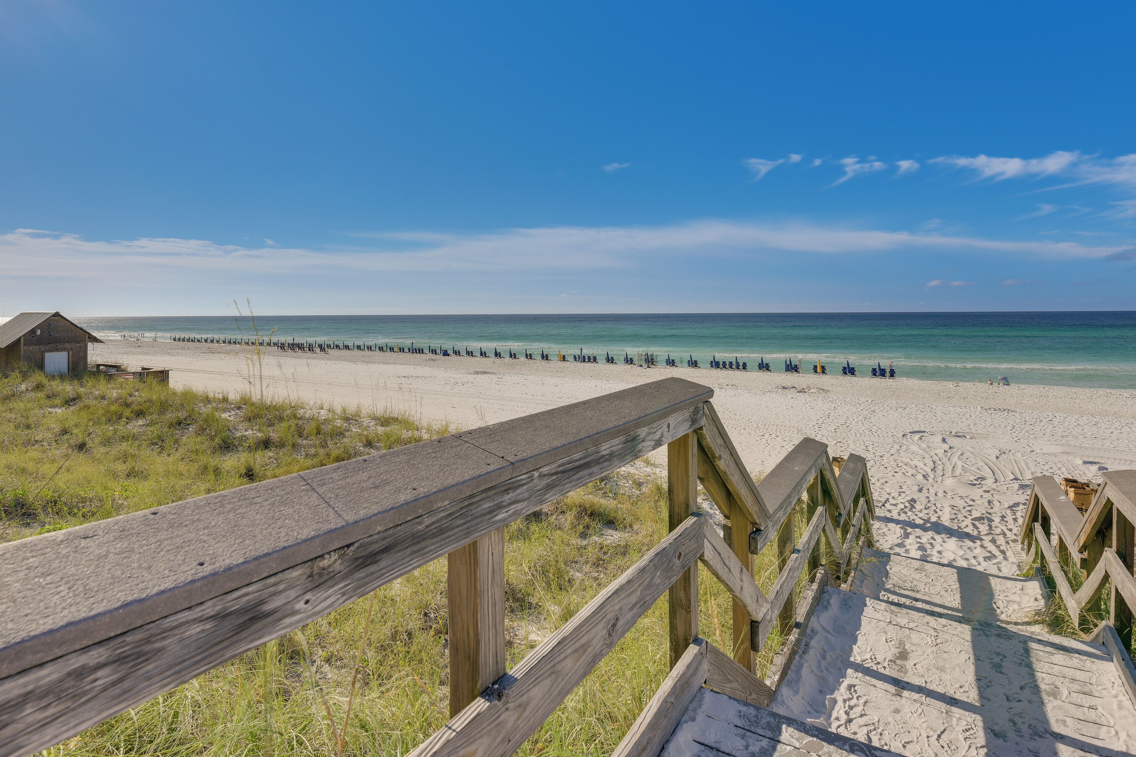 Une promenade en bois mène à une plage de sable fin, sous un ciel bleu et dégagé.