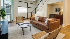 A spacious living room with a brown leather sofa, a coffee table, and a chair. A staircase leads to a second floor, and there is a large window with blinds.