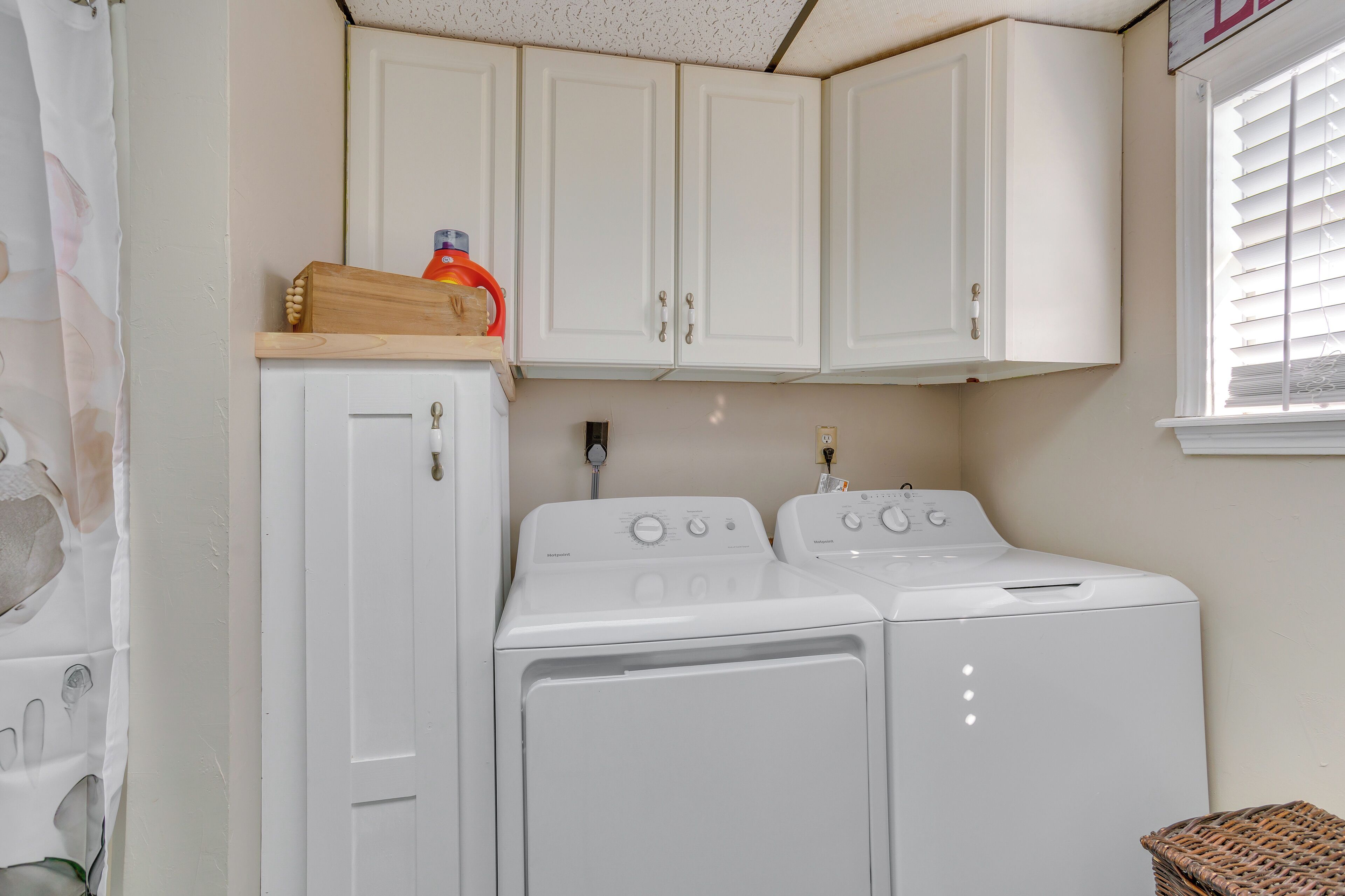 A laundry room with two white front-loading washing machines, a wooden shelf with a bottle and a container, and white cabinets above.