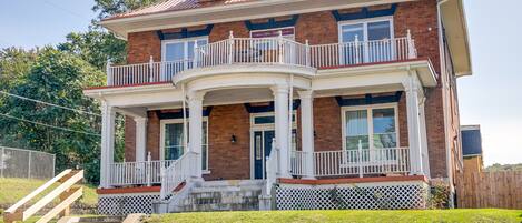 A two-story brick house with a wrap-around porch, a balcony on the second floor, and a front door with a small porch area.