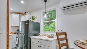 A compact kitchen with white cabinets, a built-in oven, and a refrigerator. There's a window with a view of greenery, a wooden chair, and a small dining table. - 5 Mi to Salem: Furnished Patio at Danvers Studio! (Danvers)