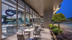 Un espacio al aire libre moderno con sillas de mimbre y mesas redondas, un letrero de Costa Coffee y el Hotel Manx al fondo.