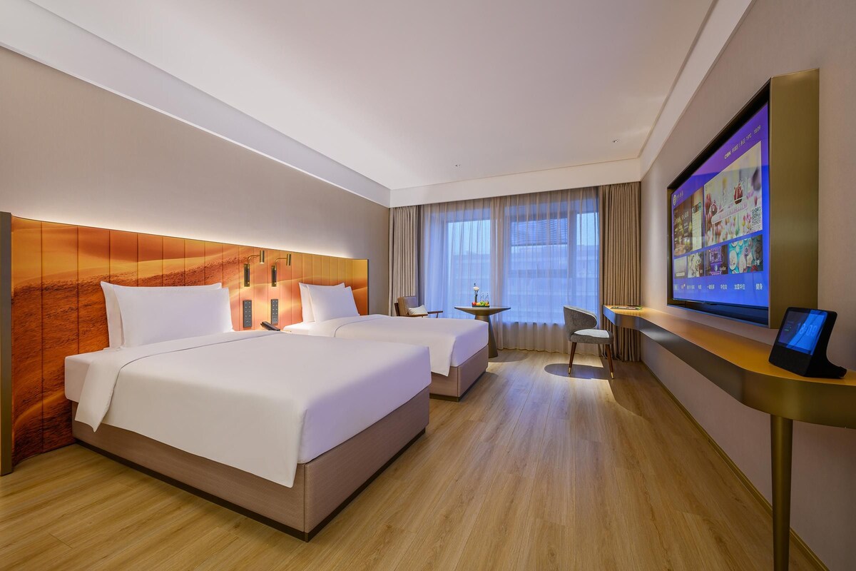A hotel room with two beds, a desk, a TV, and a large window with curtains.