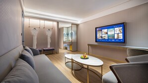 Una habitación de hotel moderna con sofá, una mesa redonda con fruta, un televisor mostrando opciones de la habitación y un espejo.