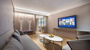 A modern hotel room with a sofa, a round table with fruit, a TV displaying room options, and a mirror.