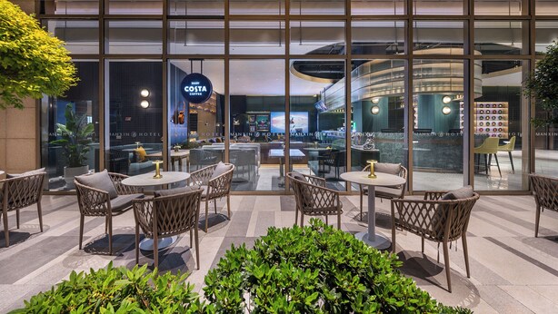 Un moderno vestíbulo de hotel con paredes de vidrio, muebles de mimbre y un letrero de Costa Coffee.