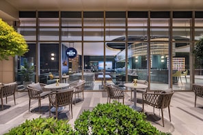 A modern hotel lobby with glass walls, wicker furniture, and a Costa Coffee sign.