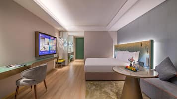 Habitación de hotel moderna con una cama, un escritorio con una silla, un televisor y una pequeña mesa con fruta y bebidas.