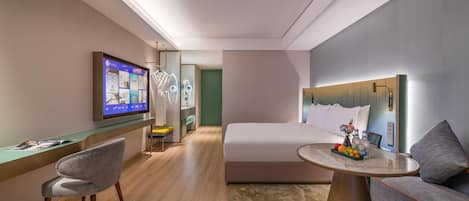 A modern hotel room with a bed, a desk with a chair, a television, and a small table with fruit and drinks.