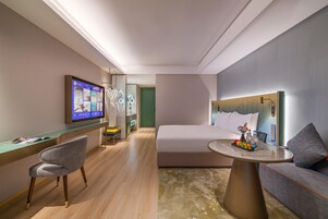 Habitación de hotel moderna con una cama, un escritorio con una silla, un televisor y una pequeña mesa con fruta y bebidas.