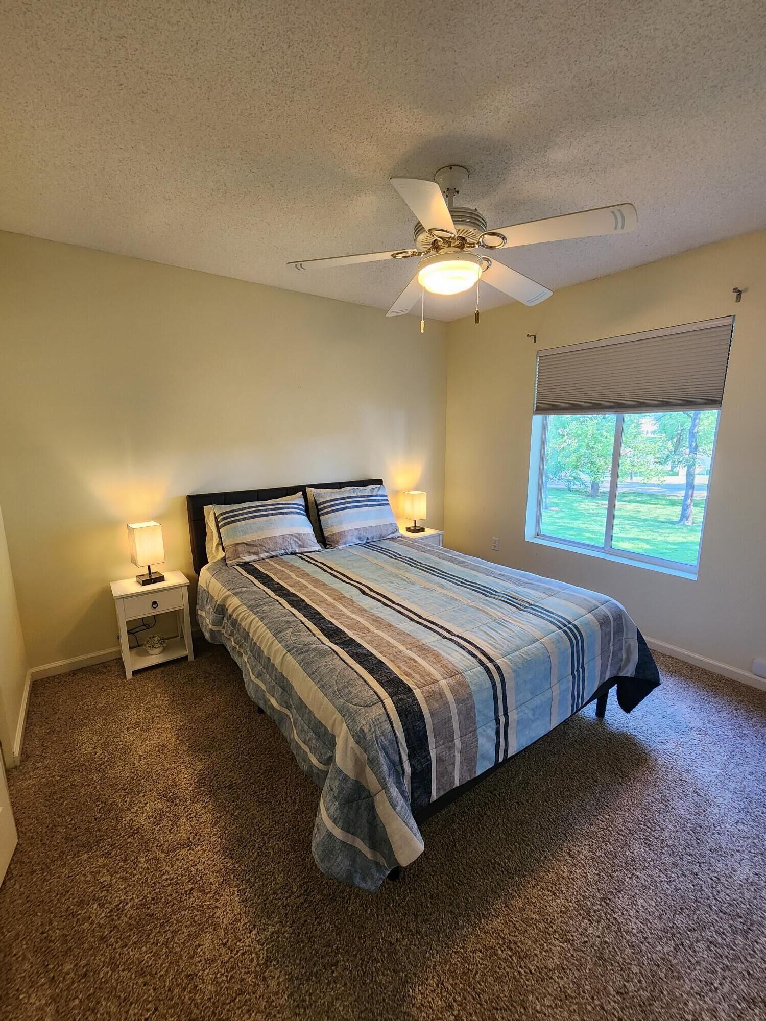 A bedroom with a bed, a ceiling fan, a window with blinds, and a nightstand with a lamp.
