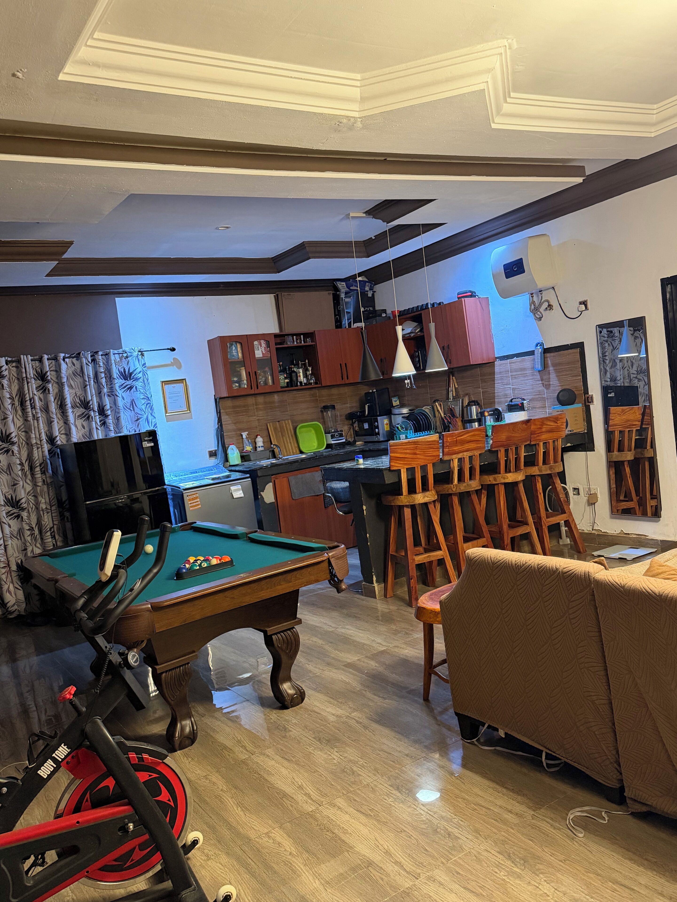 A room with a pool table, a bar area with stools, a sofa, and a kitchen in the background.