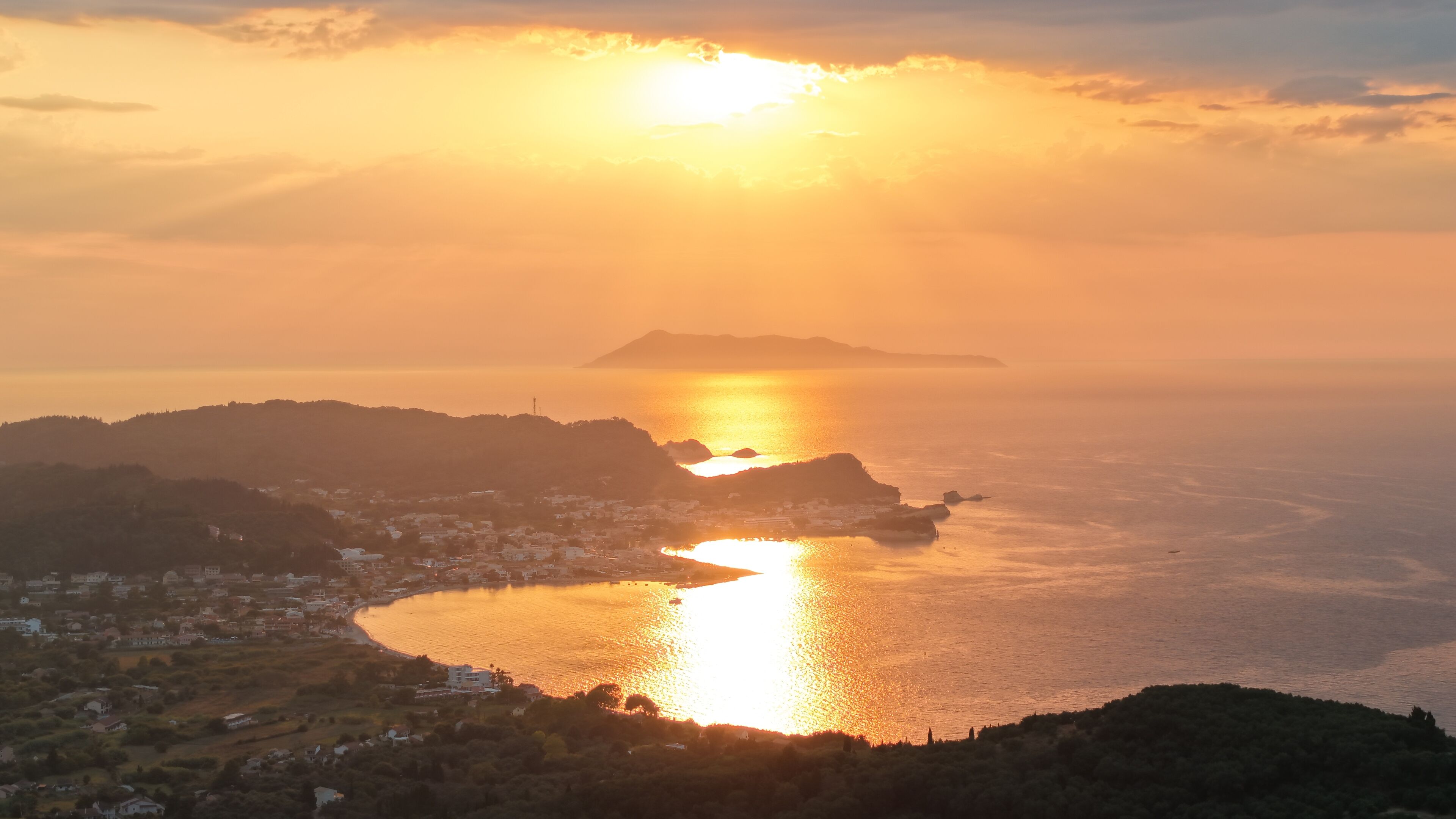 Un paysage côtier avec un coucher de soleil sur la mer, une petite île au loin et une ville avec des bâtiments le long de la côte.