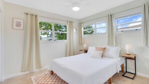 A bedroom with a bed, two bedside tables with lamps, a ceiling fan, and windows with curtains.