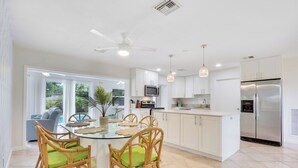 A modern kitchen with white cabinets, a stainless steel refrigerator, and a dining area with a glass table and wicker chairs.