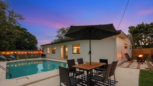 A backyard with a swimming pool, a wooden table with chairs, and a covered patio area.
